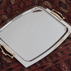 May include: A rectangular silver-toned serving tray with a polished, reflective surface and rounded corners. The tray features two ornate, gold-toned metal handles attached to opposite sides, providing a contrast to the smooth, flat center.