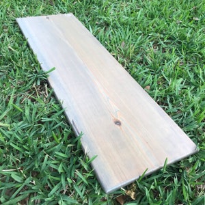 May include: A long, rectangular piece of wood with a light gray stain. The wood has a natural grain pattern and is slightly weathered.