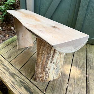 May include: A rustic wooden bench with a natural edge seat and two tree trunk-style legs. The bench is made of light-colored wood and has a smooth, flat surface. The legs are made of rough, textured wood. The bench is approximately 48 inches long.