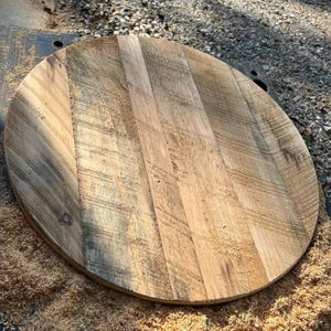 May include: A circular wooden board constructed from multiple planks of light and medium brown reclaimed wood. The surface displays visible grain patterns, natural wood textures, and saw marks, with a flat, unfinished appearance throughout.