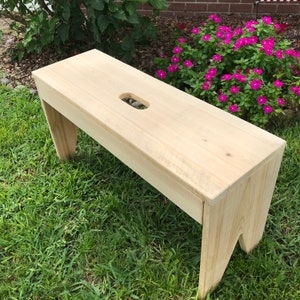 May include: A light-colored wooden bench with a rectangular top and angled legs. The bench has a handle-shaped cutout in the center of the top. The bench is outdoors on a grassy surface.