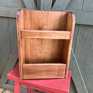 May include: A wall-mounted wooden organizer featuring a medium brown stained finish with visible wood grain. The unit has a tall back panel, two curved side supports, and a horizontal front bar positioned above a solid base shelf for storage.