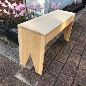 May include: A light-colored wooden bench featuring a rectangular seat and two side supports with V-shaped cutouts at the bottom. The bench is constructed from natural wood with a visible grain pattern and a smooth, light-toned finish throughout.