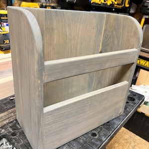 May include: A gray-stained wooden wall-mounted organizer with a curved top and two shelves. The wood grain is visible, and the piece is designed for storage. The organizer is sitting on a black surface.