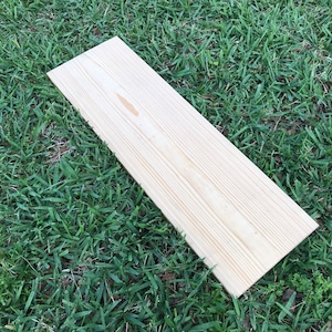 May include: A light-colored, smooth wood plank rests on a patch of vibrant green grass. The plank is rectangular, exhibiting a natural wood grain pattern. Its light beige hue suggests unfinished pine.