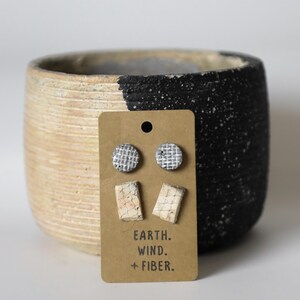 May include: A set of ceramic earrings on a brown card with the text "EARTH. WIND. + FIBER." The earrings are in two styles: a pair of small round earrings with a white and grey pattern and a pair of rectangular earrings with a white and pink pattern.