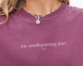 May include: A maroon t-shirt with the text "my needlepointing shirt" in white. A small illustration of scissors is below the text. A silver necklace with a circular pendant is worn around the neck.