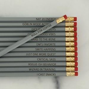 May include: A set of twelve grey pencils with gold-colored ferrules and red erasers. Each pencil features black text including phrases like NAT 20 ENERGY, GOBLIN MODE ACTIVATED, ROLL FOR COFFEE, BARD TO THE BONE, DM'S FAVORITE, SMITE HAPPENS, and I CAST SNACKS.