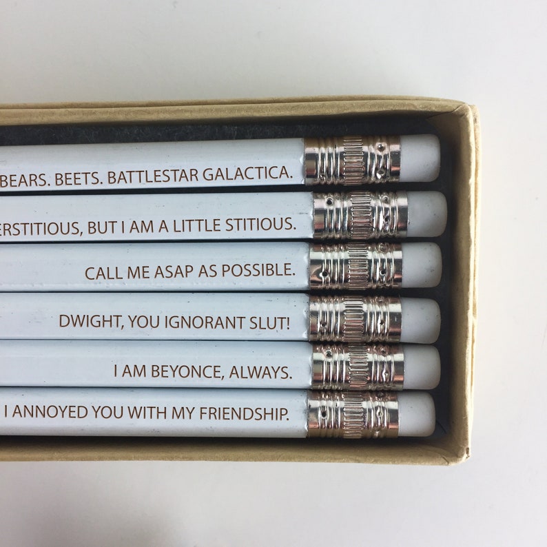 May include: A set of six white pencils with silver metal bands and erasers, each inscribed with a different quote in brown text. The quotes include phrases like "BEARS. BEETS. BATTLESTAR GALACTICA." and "DWIGHT, YOU IGNORANT SLUT!" The pencils are in a cardboard box.