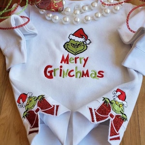 Peut inclure: Chemise à manches longues blanche avec un motif brodé de Grinch vert et rouge. La chemise dit "Merry Grinchmas" et a un Grinch avec un nœud rouge sur chaque manche.