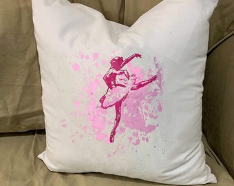 May include: A square white throw pillow featuring a centered graphic print of a ballerina in a tutu. The design is rendered in shades of pink with a distressed, splattered paint effect surrounding the figure, creating a textured visual appearance.