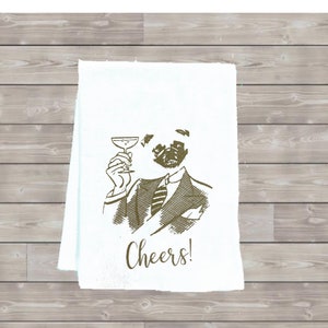 May include: A white kitchen towel with a brown illustration of a pug wearing a suit and holding a martini glass. The text "Cheers!" is printed below the illustration.