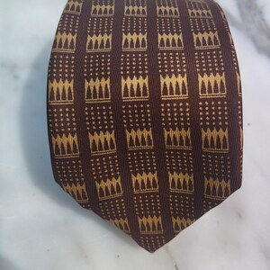 May include: A brown tie with a repeating pattern of vertical stripes and gold-colored geometric shapes. The tie is made of a textured fabric and is displayed on a white marble surface.