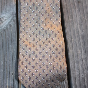 May include: A tan-colored tie with a repeating pattern of small, stylized floral designs in white, red, and blue. The tie is made of a smooth fabric and is displayed against a weathered wooden background.