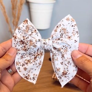 May include: A fabric hair bow featuring a white background with a repeating brown floral pattern. The bow has a structured shape with a central knot and pointed edges, showcasing a detailed print of small flowers and leaves across the surface.