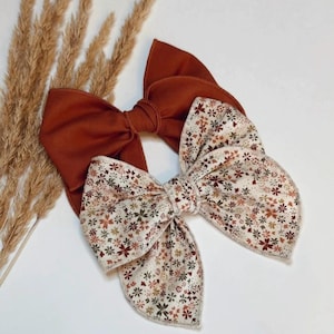 May include: Two fabric hair bows, one in a solid burnt orange color and the other in a cream fabric featuring a small-scale floral pattern with brown, green, and orange petals. Both bows have a knotted center and wide, layered fabric loops.