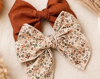 May include: Two fabric hair bows. One bow is a solid burnt orange color with a knotted center. The second bow features a cream background with a small-scale floral pattern in shades of green, orange, and brown, also with a knotted center.