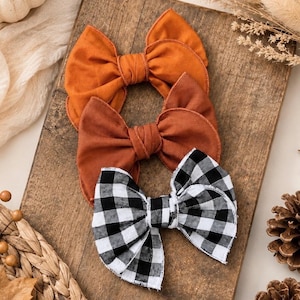 May include: Three fabric hair bows arranged vertically on a wooden board. The top bow is bright orange, the middle bow is a muted rust brown, and the bottom bow features a black and white buffalo check pattern. Each bow has a central knot detail.