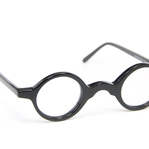 Könnte beinhalten: Schwarze, rund gerahmte Brille mit Schlüssellochsteg. Die Brille hat eine glänzende Oberfläche und dünne Bügel. Die Gläser sind klar, und der Rahmen ist einfarbig dunkel. Das Design ist klassisch und minimalistisch.