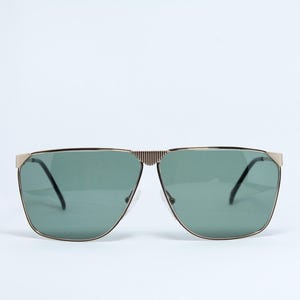 Peut inclure: Lunettes de soleil dotées d'une monture en métal doré avec une texture nervurée distincte sur le pont. Les verres sont d'une couleur vert sauge atténuée, et la monture comprend des accents dorés angulaires aux coins extérieurs.