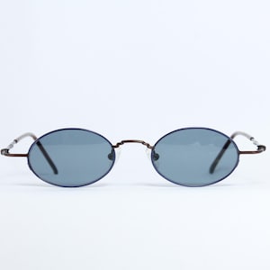 Könnte beinhalten: Ovale Sonnenbrille mit dunkelblauem Rahmen und Gläsern. Die Brille hat einen dünnen, braunen Metallsteg und Bügel. Die Gläser sind einfarbig grau. Die Sonnenbrille ist vor einem weißen Hintergrund platziert.