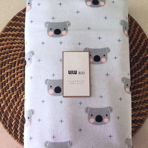 May include: A white baby blanket with a repeating pattern of gray koala bears and blue plus signs. The blanket has a tag that reads "Lilu & Co Handmade with Love".