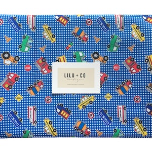 Könnte beinhalten: Blauer Stoff mit weißem Gingham-Muster, das ein sich wiederholendes Design aus verschiedenen Fahrzeugen zeigt, darunter ein Schulbus, ein Feuerwehrauto, ein Betonmischer, ein Krankenwagen, ein Taxi und ein Baufahrzeug. Der Stoff enthält auch Stoppschilder und Verkehrskegel. Der Text "LilU + Co" ist auf einem weißen Etikett gedruckt.