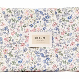 Pode incluir: Uma peça de tecido retangular com fundo branco e um padrão floral denso em tons de azul, rosa e verde. Uma etiqueta branca retangular está centrada no tecido com o texto preto LILU + CO boutique impresso nela.