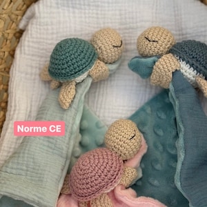 Peut inclure: Trois jouets pour bébés en forme de tortues crochetées avec des carapaces de différentes couleurs : turquoise, rose et grise. Chaque tortue est attachée à une couverture douce et texturée. Le texte « Norme CE » est visible sur l’image.
