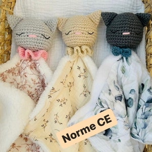 Peut inclure: Trois doudous chats au crochet avec des têtes en peluche gris clair, beige et gris foncé. Chacun présente des yeux fermés brodés, un nez rose et une collerette. Les couvertures ont des faces en tissu à motifs et des dos en peluche blanche. Texte : Norme CE.