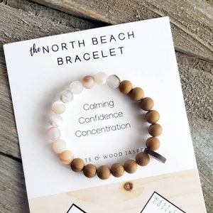 May include: A wooden beaded bracelet with a white and pink stone charm. The bracelet is on a white card with the text "the NORTH BEACH BRACELET" and "Calming Confidence Concentration TE & WOOD JASPE". The card also has a logo for "ESSENTIAL COAST" and "WEST COAST COLLECTIVE".