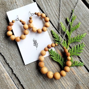 Op de afbeelding: Een set houten kralen sieraden met een ketting en oorbellen. De ketting en oorbellen zijn gemaakt van natuurlijke houten kralen en hebben een eenvoudig, elegant ontwerp. De ketting is aan een zilveren ketting bevestigd en de oorbellen hebben zilveren haakjes.