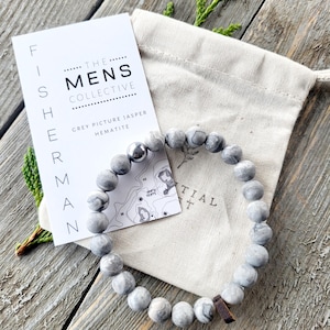 May include: A grey and white jasper and hematite beaded bracelet with a silver bead and a wooden charm. The bracelet is in a small white drawstring bag with a tag that reads "The Mens Collective" and "Grey Picture Jasper Hematite".