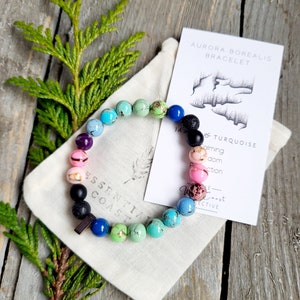 Puede incluir: Una pulsera de cuentas con una variedad de cuentas coloridas, incluyendo azules, verdes, rosas y negras, descansa sobre una pequeña bolsa de lona. Una tarjeta con el texto "Aurora Borealis Bracelet" y un dibujo de un paisaje está junto a la pulsera. El fondo es una superficie de madera.