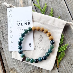 May include: A green and brown beaded bracelet with a wooden bead accent. The bracelet is in a small white drawstring bag with the text "The Mens Collective" and "Essential Coast" printed on a card.
