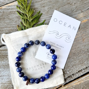 Könnte beinhalten: Ein blaues Stein-Armband mit silbernen Perlen und einem Holz-Anhänger. Das Armband befindet sich in einem weißen Stoffbeutel mit einer Karte, auf der "Ocean, Peace, Balance & Serenity" steht.
