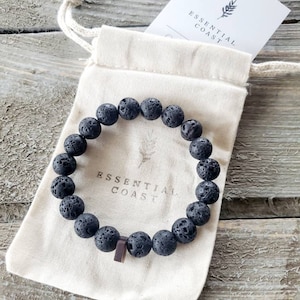 May include: A black lava rock bracelet with a wooden bead accent. The bracelet is in a white drawstring bag with the text "Essential Coast" printed on it.