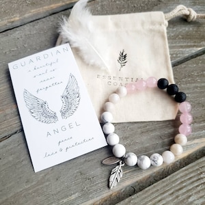 May include: A white and pink beaded bracelet with a silver feather charm. The bracelet is made with white howlite, pink rose quartz, and black lava beads. The card reads "Guardian Angel, a beautiful soul is never forgotten, peace, love & protection".