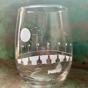 May include: Clear stemless wine glass with etched design. The design includes a sun, birds, a bridge, and a boat on water. The text "Call me when you get to the" is etched on the bottom of the glass.