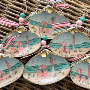 May include: Hand-painted seashell ornaments with a beach scene featuring a lighthouse, beach chairs, and an umbrella. Each shell is adorned with a gold rim, a ribbon, and beads. The text "BEACH HAVEN 2026" is written on each shell.
