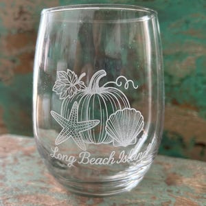 May include: A clear stemless wine glass with a white etched design. The design includes a pumpkin, starfish, seashell, and the words "Long Beach Island". The glass is perfect for serving wine.