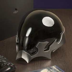 May include: A black helmet with a silver, angular design. The helmet has a glossy finish and is sitting on a wooden surface.