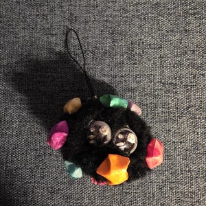 May include: A black, fluffy ornament with large, clear, round eyes and an orange beak. The ornament is decorated with colourful, gem-like accents in pink, green, and yellow. A black string is attached for hanging.