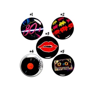 May include: Five round buttons with different 80s themed designs. The buttons are numbered 1 through 5. Button 1 has a neon pink and white "80's" graphic on a black background with blue and white lines. Button 2 has a black background with a repeating pattern of yellow and pink space invaders. Button 3 has a red lip print on a black background. Button 4 has a black vinyl record with a red center on a white background. Button 5 has a cassette tape with a rainbow design on a black background.