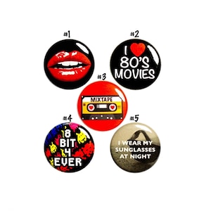 May include: Five 1980s themed buttons. The first button has a red pair of lips on a black background. The second button has a red heart with the text "I ♥ 80's MOVIES" on a black background. The third button has a red cassette tape with the text "MIXTAPE" on a yellow background. The fourth button has a colorful pixelated design with the text "8 BIT 4 EVER". The fifth button has a black and white image of a person wearing sunglasses at night with the text "I WEAR MY SUNGLASSES AT NIGHT".