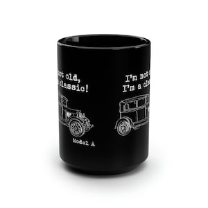 May include: Black ceramic mug featuring white line drawings of vintage Model A cars. The mug has the text "I'm not old, I'm a classic!" and "Model A" printed on it. The mug is a cylindrical shape.