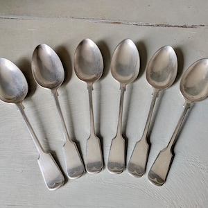 May include: Six antique silver spoons arranged in a fan shape. Each spoon has an oval bowl and a long, slender handle with a decorative end. The spoons are displayed on a light gray surface.