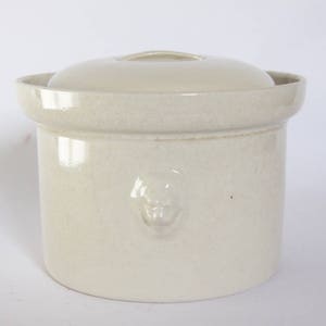 以下が含まれることがあります： 蓋付きの白い陶器の壺。壺は表面がざらざらしていて、前面に小さな装飾的なデザインが施されています。
