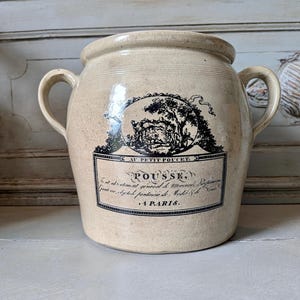 Può includere: Un vaso in ceramica beige con due manici laterali e una stampa nera a trasferimento raffigurante un'illustrazione paesaggistica, il testo AU PETIT POUCET, POUSSE e A PARIS, insieme a ulteriore piccolo testo stampato entro un bordo rettangolare.