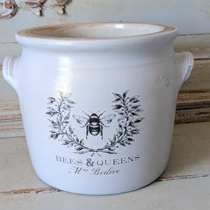 Könnte beinhalten: Ein weißer Keramiktopf mit zwei seitlichen Griffen, verziert mit einer schwarzen Grafik einer Biene in einem Blattkranz. Unter dem Kranz steht der Text BEES & QUEENS und Mme Boileve. Der innere Rand ist hellbraun und die Außenseite ist matt gehalten.